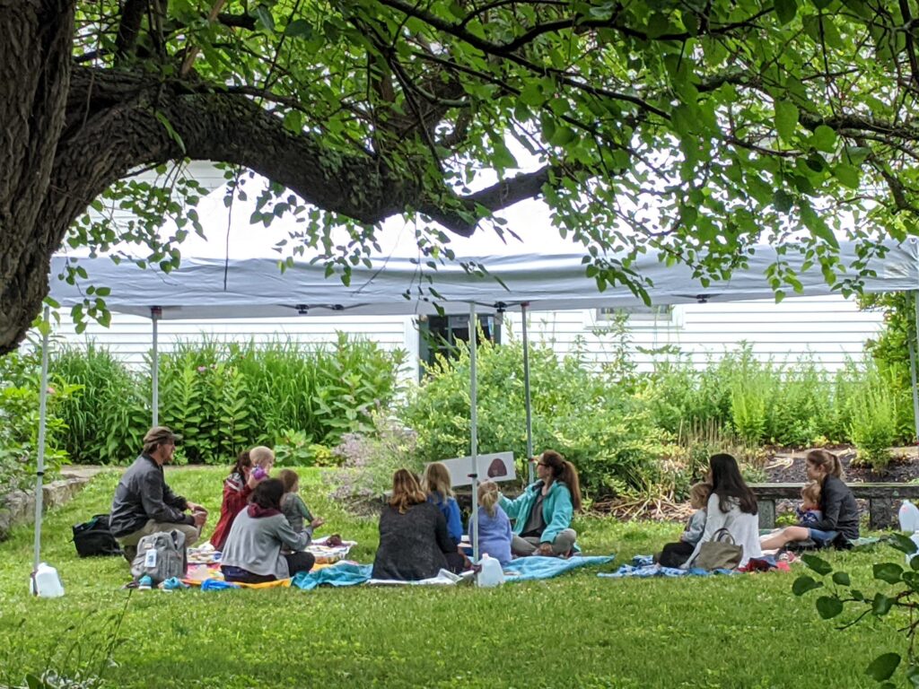 Woman reading aloud to a group of families under a tent canopy
